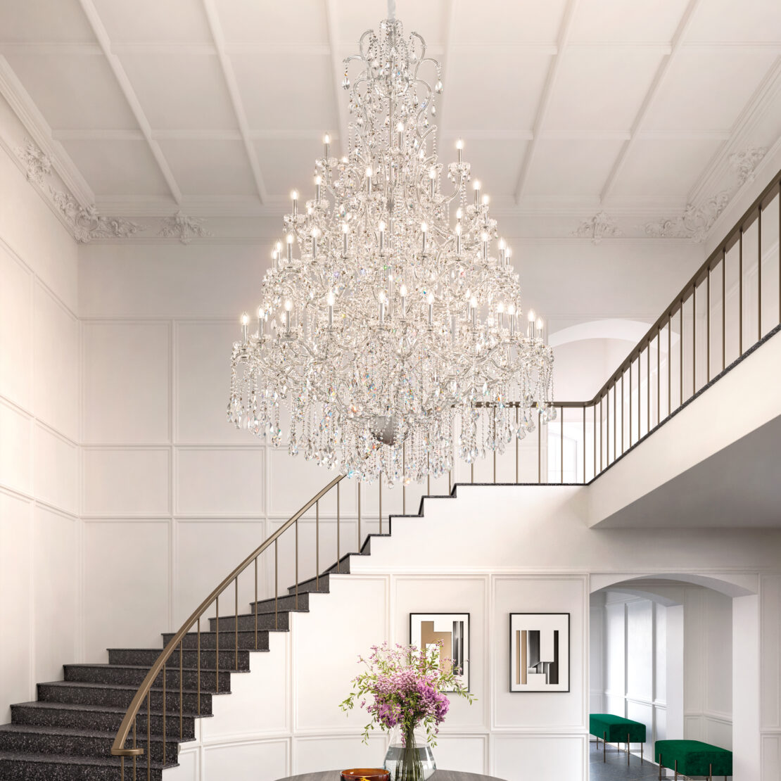 A Grand Crystal Crystal Chandelier hangs in a hallway above a spiral staircase in an elegant foyer with white walls, black railings, framed art, and a round flower-topped table. Green chairs are seen in the background.