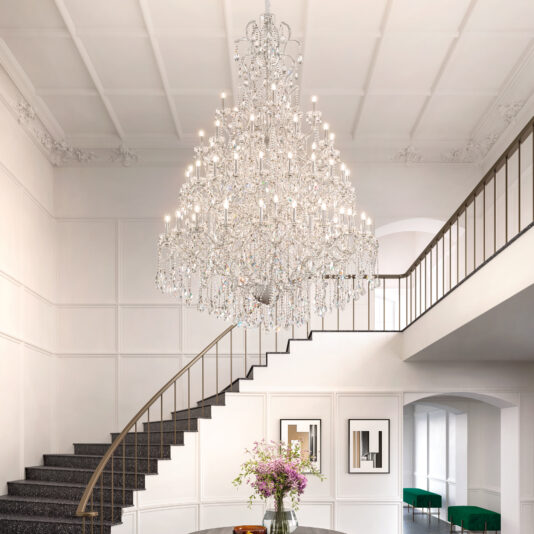 Large Statement Crystal Chandelier A Grand Crystal Crystal Chandelier hangs above a spiral staircase in an elegant foyer with white walls, black railings, framed art, and a round flower-topped table. Green chairs are seen in the background.