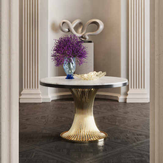 The Round Marble Contemporary Dining Table features a gold fluted base and white marble top, set on a dark patterned floor. A blue vase with purple flowers and a golden decorative piece adorn the table, with sculptural art displayed behind.