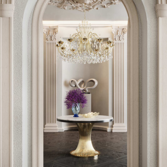 Round Marble Contemporary Dining Table A luxurious interior featuring the Round Marble Contemporary Dining Table with a gold base, an ornate chandelier, purple flowers in a vase, intricate wall moldings, and a modern silver sculpture in the background.
