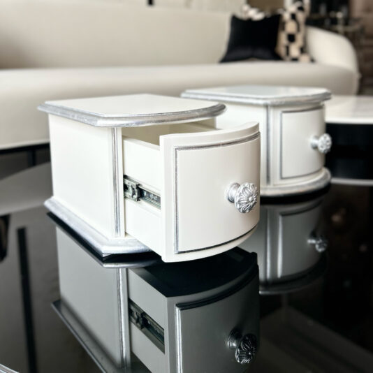 Two small, elegant Classic Style 2 Drawer Bedside Cabinets with silver trim and ornate knobs rest on a reflective black glass table; one drawer is partially open. A modern white sofa is visible in the background. The concepts for these pieces of furniture were design using AI