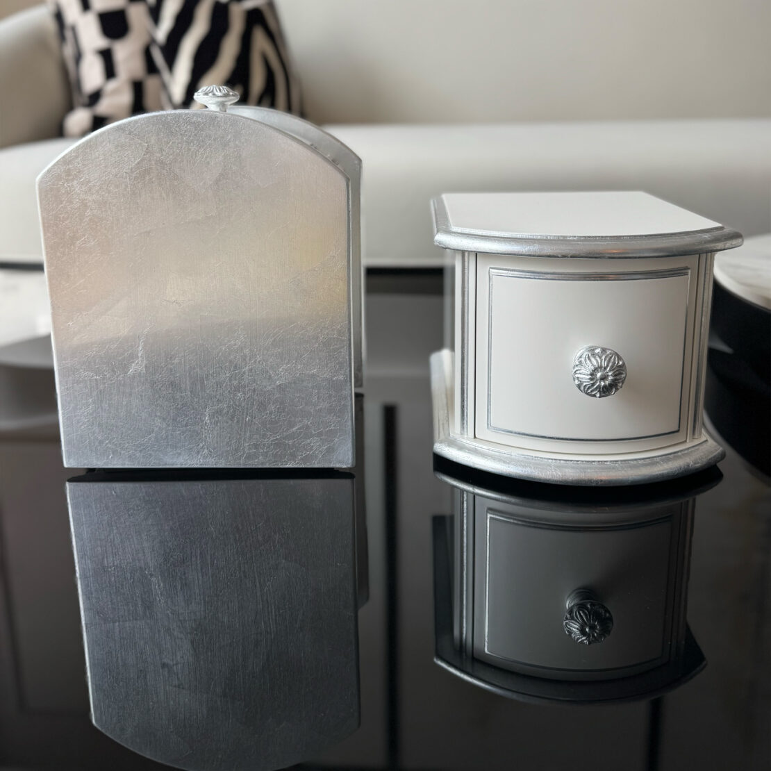 Two small decorative boxes, inspired by the Classic Style 2 Drawer Bedside Cabinet (Copy), one silver and one white with silver trim and ornate knobs, rest on a glossy black surface, reflecting beside a patterned pillow and white couch in the background.