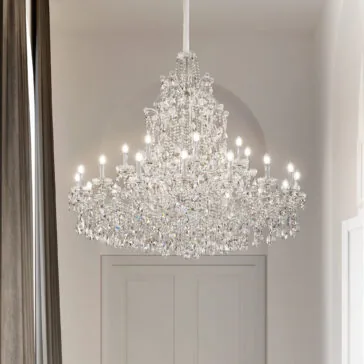 Tiered Clear Crystal Chandelier A Tiered Clear Crystal Chandelier with glowing bulbs hangs from the ceiling in a bright, elegant space with high ceilings, white walls, and tall dark curtains.