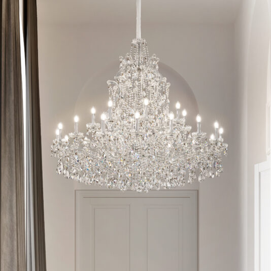A Tiered Clear Crystal Chandelier with glowing bulbs hangs from the ceiling in a bright, elegant space with high ceilings, white walls, and tall dark curtains.