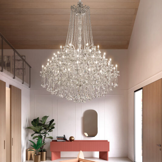 The Grand Tiered Crystal Empire Chandelier (Copy) hangs from a wooden ceiling in a bright modern room with a pink console table, oval mirror, indoor plant, and wooden accents. Sunlight pours in through a tall doorway.