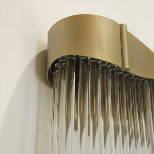 Close-up of the Contemporary Wavy Tiered Chandelier, a wall-mounted fixture with a brushed gold frame and cascading vertical glass rods, set against a white wall.