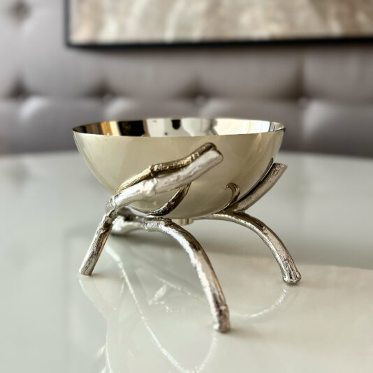 The Nut Bowl On Branch Design features a shiny silver bowl resting on a sculpted metal stand with three branch-like legs, displayed on a glossy white surface against a softly blurred background.