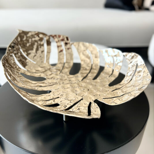 Tropical Leaf Basket The Tropical Leaf Basket, featuring a monstera shape and shiny gold hammered finish, adds elegance atop a round black table, with a blurred white sofa in the background of this stylish scene.