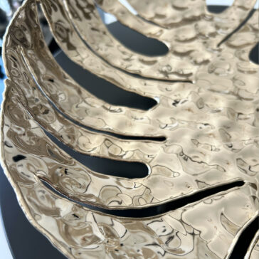 A close-up of the Tropical Leaf Basket shows its metallic gold finish, hammered texture, and cutout design resembling a stylized leaf or fan. The shiny surface reflects light for a glossy effect.
