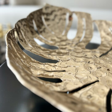 A close-up of the Tropical Leaf Basket showcases its metallic finish, hammered texture, multiple cutouts, and shiny reflective surface against a softly blurred background.