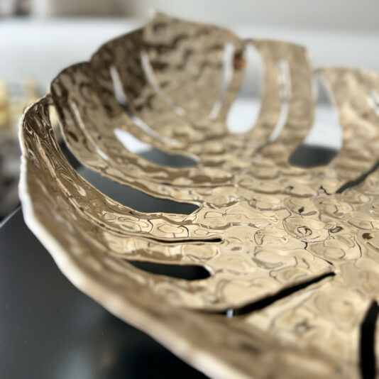 Tropical Leaf Basket A close-up of the Tropical Leaf Basket showcases its metallic finish, hammered texture, multiple cutouts, and shiny reflective surface against a softly blurred background.