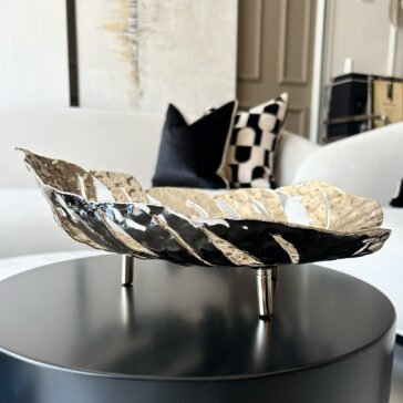 The Tropical Leaf Basket, featuring a modern metallic leaf design, is displayed on a black round table in a chic living room with a white sofa and monochrome patterned cushions.