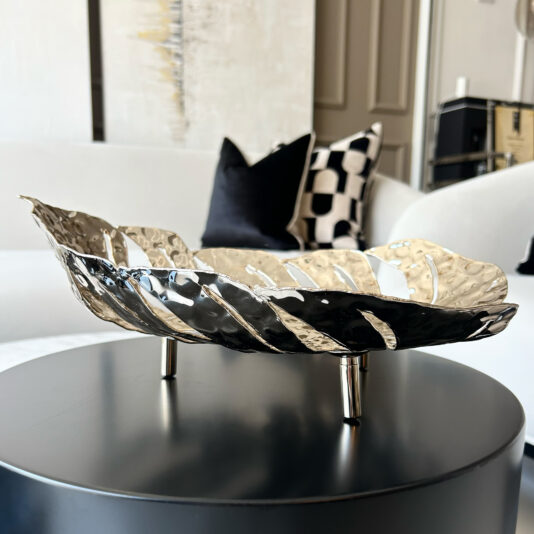 Tropical Leaf Basket The Tropical Leaf Basket, featuring a modern metallic leaf design, is displayed on a black round table in a chic living room with a white sofa and monochrome patterned cushions.