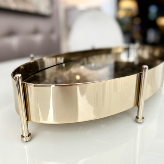 A Tropical Leaf Oval Tray With Feet, gold-colored with four vertical supports, is displayed close-up on a glossy white surface against a blurred backdrop.