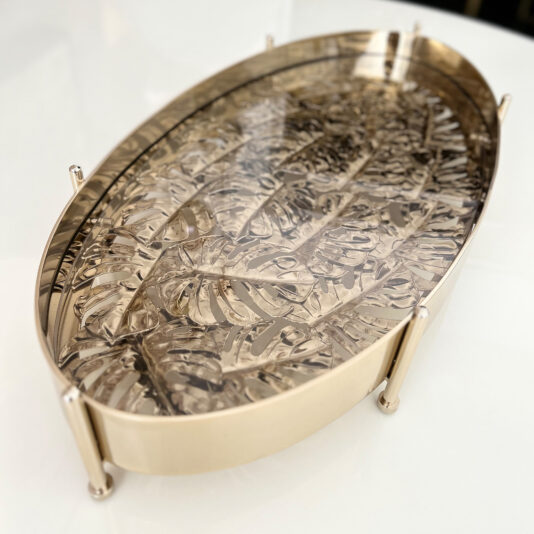 The Tropical Leaf Oval Tray With Feet boasts a shiny gold finish, raised edges, and an intricate leaf pattern, adding elegance and sophistication to any white surface.