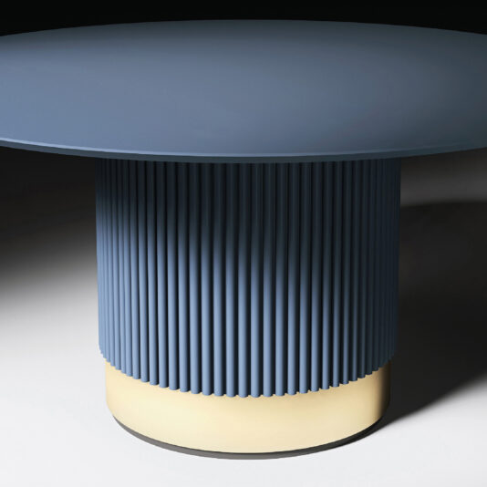 The Round Contemporary Lacquered Dining Table features a smooth blue top and cylindrical, vertically grooved base on a yellow circular platform. Dramatic lighting highlights its modern design, casting striking shadows.