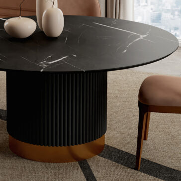 The Round Contemporary Lacquered Dining Table with white veining and a gold fluted base is paired with a tan upholstered chair. Two white vases sit on the table, set before a blurred cityscape background.