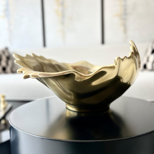 The Decorative Wave Ornament (Copy), a gold abstract bowl with wavy edges, rests on a round black table in a modern, bright living room with blurred couches and checkered pillows in the background.