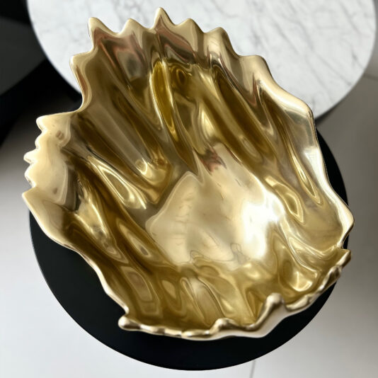 Luxury Decorative Wave Bowl The Decorative Wave Ornament (Copy), a shiny gold, clam-shaped bowl with wavy edges, sits on a round black table, with part of a white marble table visible in the background.