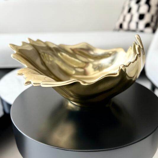 Luxury Decorative Wave Bowl A gold, abstract, leaf-shaped decorative bowl rests on a round black table with the Decorative Wave Ornament (Copy), while a white sofa and patterned cushion appear blurred in the background.