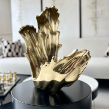 A shiny gold abstract sculpture sits on a black round table beside a Set Of 3 Luxury Marble Candle Holders (Copy) in a modern living room with a white sofa, patterned pillows, and a chessboard in the background.