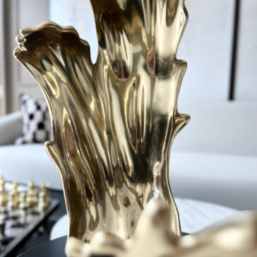 A close-up of a shiny, abstract gold sculpture with organic curves. In the blurred background, a sofa and chessboard are visible, complemented by a Set Of 3 Luxury Marble Candle Holders (Copy) that adds elegance to the decor.