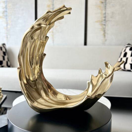A modern gold sculpture with fluid curves sits on a round black table next to a Set Of 3 Luxury Marble Candle Holders (Copy), with a light sofa and patterned cushions softly blurred in the background.