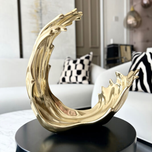 Luxury Decorative Wave Ornament A shiny gold abstract wave sculpture sits on a black round table next to a Set Of 3 Luxury Marble Candle Holders (Copy), all in a modern living room with neutral tones and geometric decor.