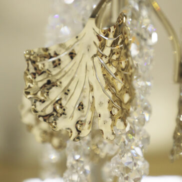 A close-up of a shiny gold leaf-shaped ornament with detailed ridges from the Contemporary Leaf Chandelier With Crystal Drops (Copy), surrounded by dangling faceted clear crystals that reflect light.