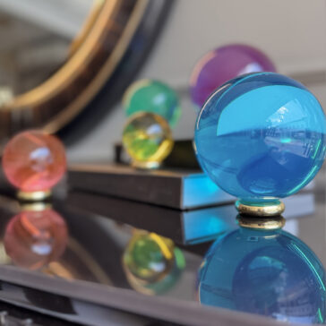 Luxury Colourful Acrylic Ball Set The Luxury Chess Set (Copy) features colorful glass spheres in various sizes, with the largest in blue and others in purple, green, yellow, and red. Arranged on a reflective surface before a mirror, they evoke refined elegance amid blurred books.