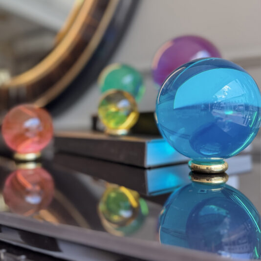 The Luxury Chess Set (Copy) features colorful glass spheres in various sizes, with the largest in blue and others in purple, green, yellow, and red. Arranged on a reflective surface before a mirror, they evoke refined elegance amid blurred books.