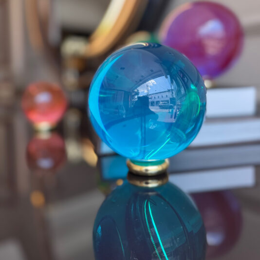 A close-up of a blue glass sphere from the Luxury Chess Set (Copy) sits on a reflective surface, with pink and orange spheres blurred in the background and vivid reflections highlighting its elegance.