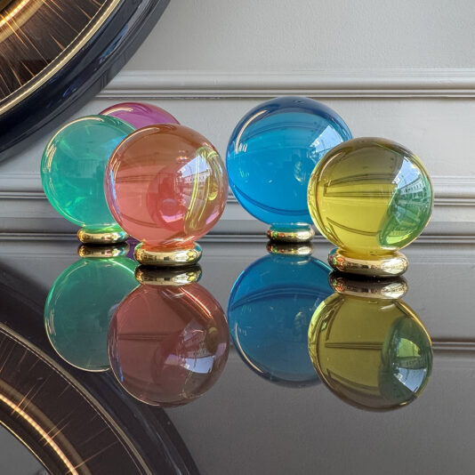 The Luxury Chess Set features five vibrant glass orbs—teal, pink, orange, blue, and yellow—on gold bases, resembling chess pieces reflected on a glossy black surface against a white wall with decorative molding.