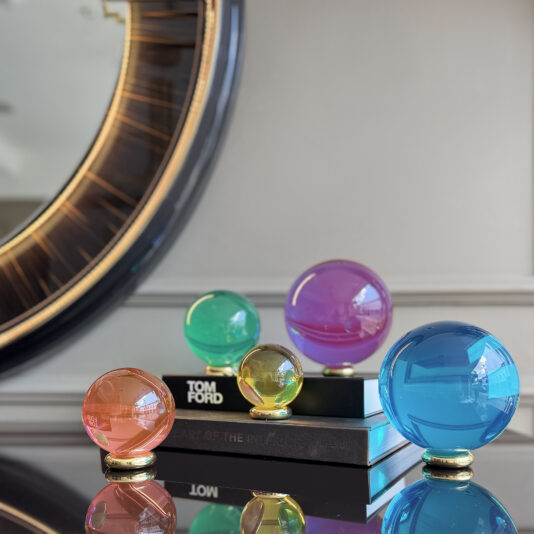 The Luxury Chess Set (Copy), with five colorful glass spheres on stands atop two stacked books before a gold-trimmed mirror, brings elegance and sophistication against a gray paneled wall.