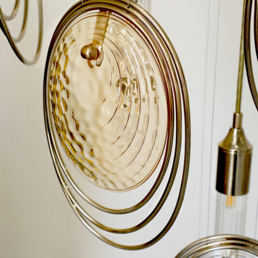 Modern Ceiling Light Inspired By The Sea Close-up of the Modern Ceiling Light Inspired By The Sea, showing concentric metallic rings and a textured, warm-tinted circular glass detail; a cylindrical metallic lamp appears in the background.