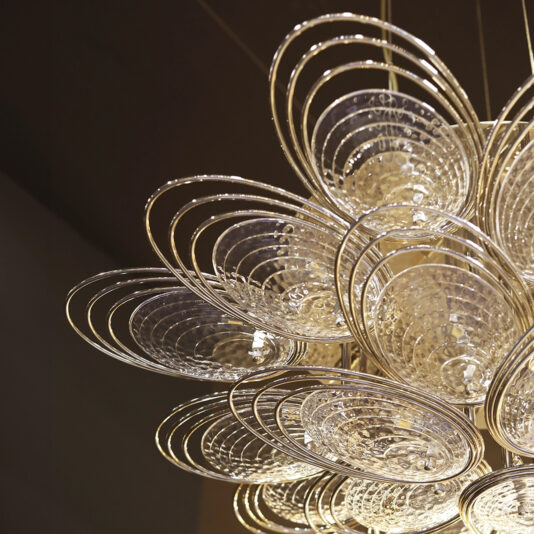 Modern Ceiling Light Inspired By The Sea The Modern Stairwell Chandelier Inspired By The Sea (Copy) features layered circular glass and gold metal elements in a floral design, reflecting warm light for an elegant glow.