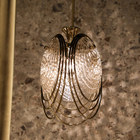 Modern Pendant Light Inspired By The Sea The Modern Round Chandelier Inspired By The Sea features a textured glass bulb encased in decorative gold metal rings and hangs before a speckled gray stone wall, casting a warm glow.