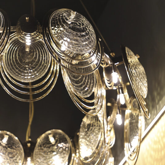 Modern Round Chandelier Inspired By The Sea Close-up of the Large Leaf Chandelier With Crystal Drops (Copy), showcasing circular, textured glass and metal fixtures with crystal drops that cast a warm, ambient light against a dark background.