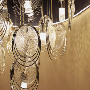Modern Stairwell Chandelier Inspired By The Sea The Modern Round Chandelier Inspired By The Sea (Copy) features circular, textured glass and metal rings that cast intricate reflections and shadows on a beige wall.