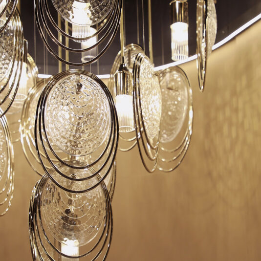 The Modern Round Chandelier Inspired By The Sea (Copy) features circular, textured glass and metal rings that cast intricate reflections and shadows on a beige wall.