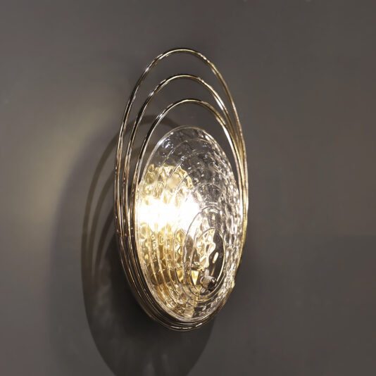 Modern Wall Light Inspired By The Sea The Modern Ceiling Light Inspired By The Sea features a textured transparent glass dome and three gold metal rings, casting a warm glow against a gray wall.