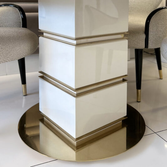 The Mother Of Pearl Exclusive Round Dining Set features a glossy white square pedestal with gold trim atop a round, reflective gold base, paired with upholstered chairs with black legs, all set on a shiny white floor.