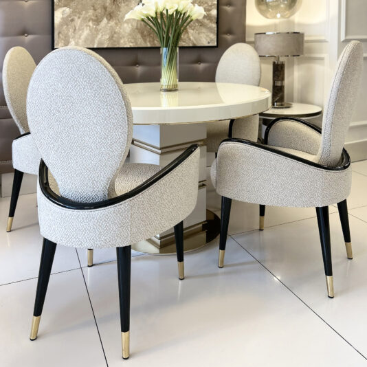 A modern dining area with the Mother Of Pearl Exclusive Round Dining Set: a round white table and four rounded-back, upholstered chairs with black legs and gold tips, arranged around a vase of white flowers.