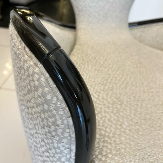 Close-up of a modern upholstered chair with textured light fabric and shiny black curved armrest from the Mother Of Pearl Exclusive Round Dining Set, shown on a white tiled floor.