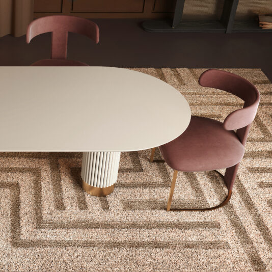 Oval Contemporary Lacquered Dining Table The Oval Contemporary Lacquered Dining Table with a rounded white top and fluted base is paired with two mauve velvet chairs on a beige geometric rug, creating a minimalist and stylish setting.