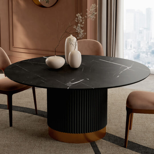 A modern dining room showcases a Round Contemporary Lacquered Dining Table (Copy) with a gold base, surrounded by tan upholstered chairs and white vases with dried branches. Floor-to-ceiling windows reveal a cityscape backdrop.