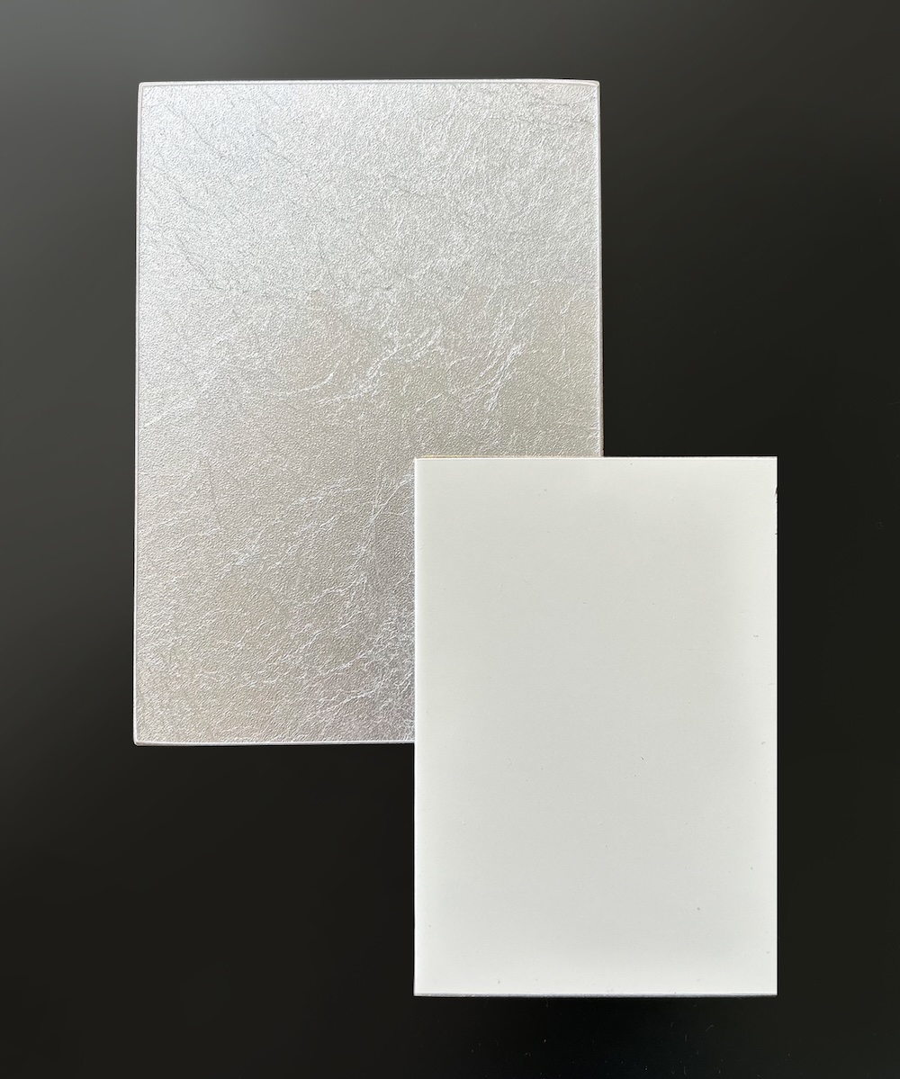 Two rectangular materials on a black sample board: one larger, textured and metallic silver, overlapping a smaller, smooth, matte white rectangle sent to our client for approval of materials