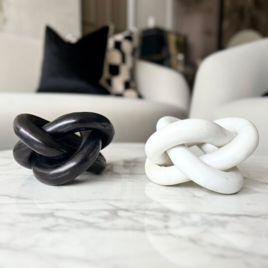 Set Of Luxury Marble Knot Ornaments Two intertwined knot sculptures, one black and one white, are displayed on a marble table with a Set of 3 Burr Walnut Effect Photo Frames (Copy) in a modern living room featuring white armchairs and a black pillow in the background.