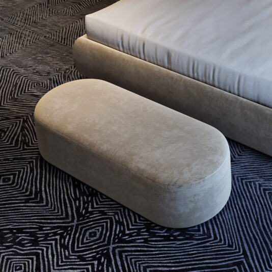 A Contemporary Curved Oval Bench in beige sits at the foot of a bed with a white mattress, atop a dark geometric-patterned carpet.