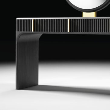 A close-up of the Contemporary Lacquered Dressing Table in black, showcasing vertical grooves, gold accents, one visible leg, and part of a round mirror against a white and black background.